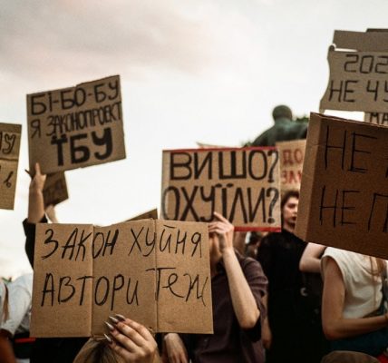 Protesters hold signs with handwritten messages against a cloudy sky.