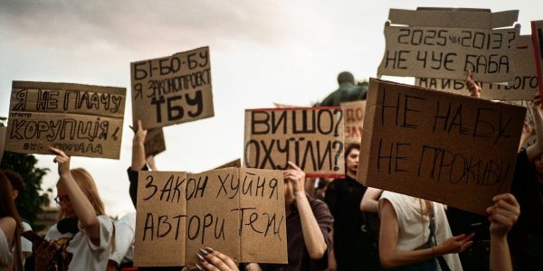 Protesters hold signs with handwritten messages against a cloudy sky.